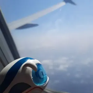 Figur Benni IC Bus guckt aus dem Fenster eines Flugzeugs auf Wolken.