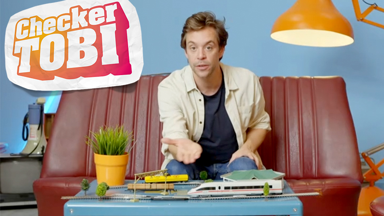 Checker TOBI sitzt auf einer brauner Sofa und moderiert eine Sendung zum Thema Bahnhofs-Check!