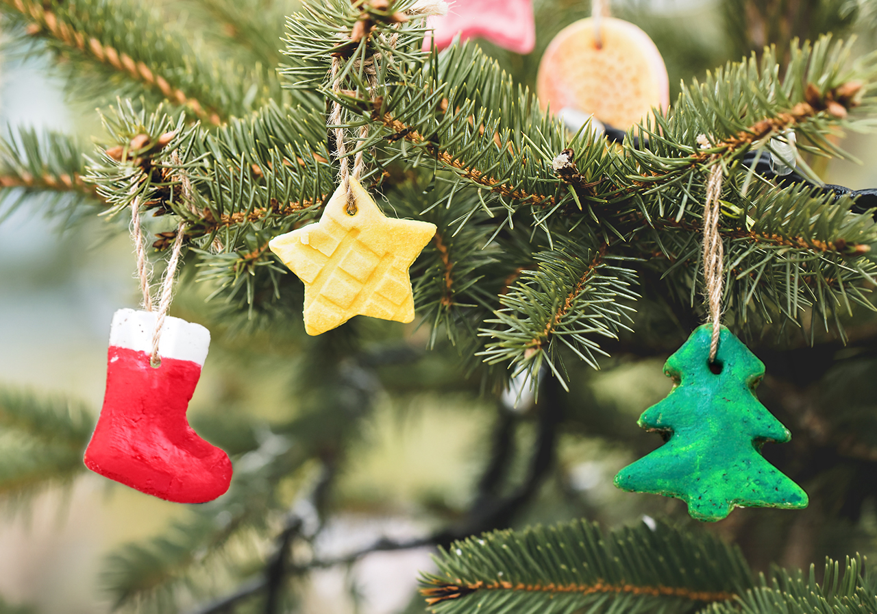 Weihnachtsschmuck aus Salzteig – ein roter Stiefel, ein gelber Stern und ein grüner Tannenbaum – hängt an den Zweigen eines Weihnachtsbaums.