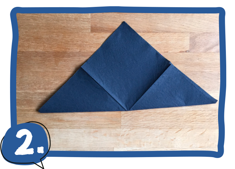 Eine dunkelblaue Serviette liegt als großes Dreieck gefaltet auf einem Holztisch, die untere Spitze wurde nach oben geklappt. Schritt 2 der Bastelanleitung.