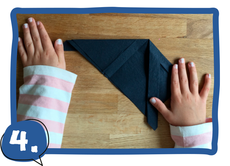 Zwei Kinderhände legen eine dunkelblaue, gefaltete Serviette schräg auf einen Holztisch, die rechte Ecke ist nach unten geklappt. Schritt 4 der Bastelanleitung.