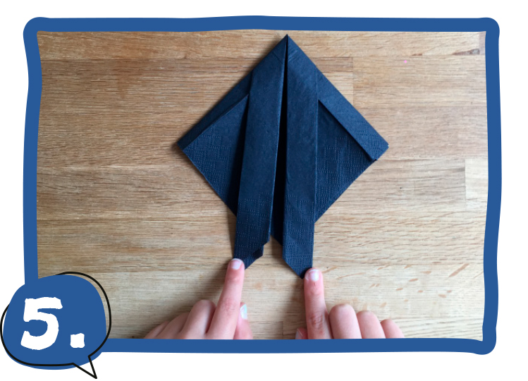 Zwei Kinderfinger zeigen auf die untere Spitze einer dunkelblau gefalteten Serviette, die eine rautenähnliche Form mit zwei Zipfeln unten hat. Schritt 5 der Bastelanleitung.