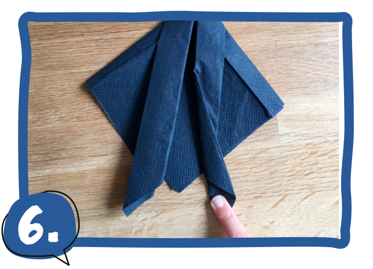 Ein Kinderfinger hält die untere Spitze einer dunkelblau gefalteten Serviette, die nun aufgefächerte Lagen und zwei seitliche Zipfel als Schwanzflosse zeigt. Schritt 6 der Bastelanleitung.