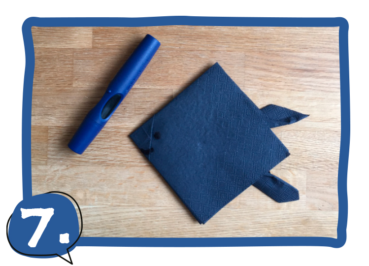 Eine dunkelblaue gefaltete Serviette in Form eines Fisches mit zwei Flossen liegt neben einem blauen Stift auf einem Holztisch. Schritt 7 der Bastelanleitung.Sonnet 4.6