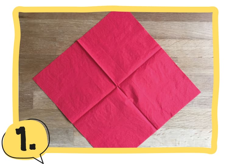 Eine rote Serviette liegt aufgefaltet als Quadrat auf einem Holztisch, mit der Ecke nach oben ausgerichtet. Schritt 1 der Bastelanleitung.