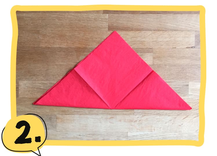 Eine rote Serviette liegt als großes Dreieck auf einem Holztisch, die untere Spitze wurde nach oben auf die obere Spitze gefaltet. Schritt 2 der Bastelanleitung.