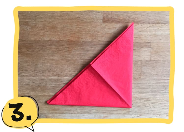 Eine rote Serviette liegt als Dreieck gefaltet auf einem Holztisch, die rechte Ecke wurde auf die linke geklappt. Schritt 3 der Bastelanleitung.