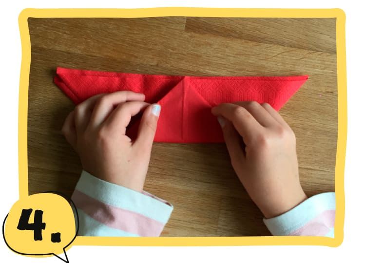 Zwei Kinderhände halten eine gefaltete rote Serviette in Dreiecksform und knicken die untere Spitze nach oben. Schritt 4 der Bastelanleitung.