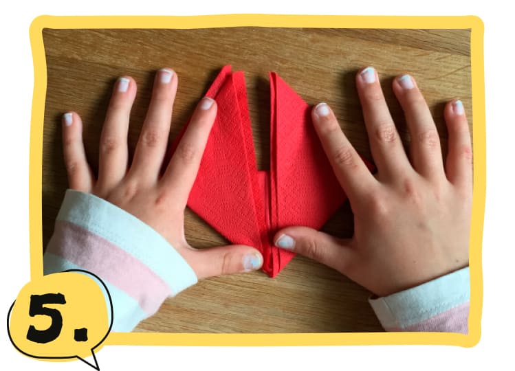 Zwei Kinderhände falten eine rote Serviette auf einem Holztisch, die rechte und linke Ecke werden nach oben geklappt. Schritt 5 der Bastelanleitung.