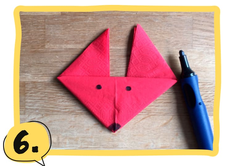 Gefaltete rote Serviette in Form eines Fuchskopfes mit zwei schwarzen Punkten als Augen, neben einem blauen Stift auf einem Holztisch. Schritt 6 der Bastelanleitung.