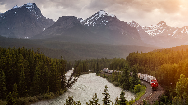 Ein langer Zug fährt an einem Fluss durch einen Wald; dahinter hohe, schneebedeckte Berge.