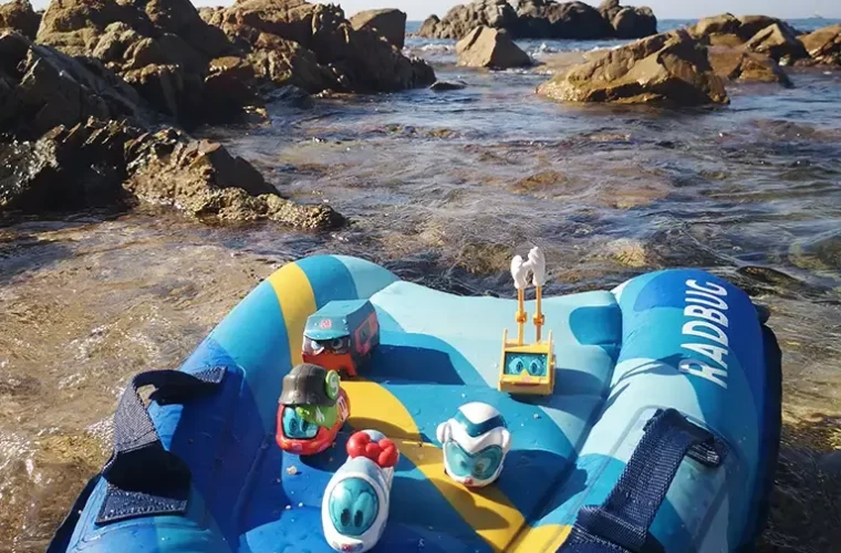 Fünf Spielfiguren stehen auf einer Luftmatratze am Strand vor Felsen.