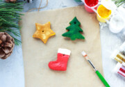 Bild 4 – Anmalen und Verzieren Bemalte Salzteigfiguren – Stern, Tannenbaum und Stiefel – liegen auf Backpapier neben Farben und einem Pinsel.