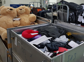 Zwei große Teddybären sitzen in einer Metallkiste. Daneben liegen Kleidung und ein rotes Reisekissen in einer zweiten Kiste.
