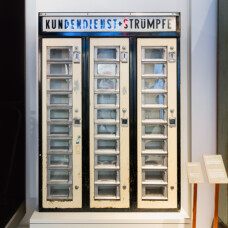 Historischer Strumpfautomat mit mehreren Fächern und Beschriftung „Kundendienst Strümpfe“ in einer Ausstellung.