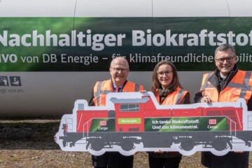 Auch Güter- und Rangierloks der DB tanken bereits umweltfreundlichen Kraftstoff. Dafür wurde z.B. eine Tankstelle am Rangierbahnhof München-Nord wurde dafür komplett auf den Biokraftstoff HVO (Hydrotreated Vegetable Oil) umgestellt. Sigrid Nikutta ist Vorstand für den Güterverkehr der Deutschen Bahn und freut sich bei der Eröffnung mit ihren Kollegen über die neue Biokraft-Tankstelle. Drei Menschen in orangenen Westen stehen vor einem Tank mit der Aufschrift „Nachhaltiger Biokraftstoff“.